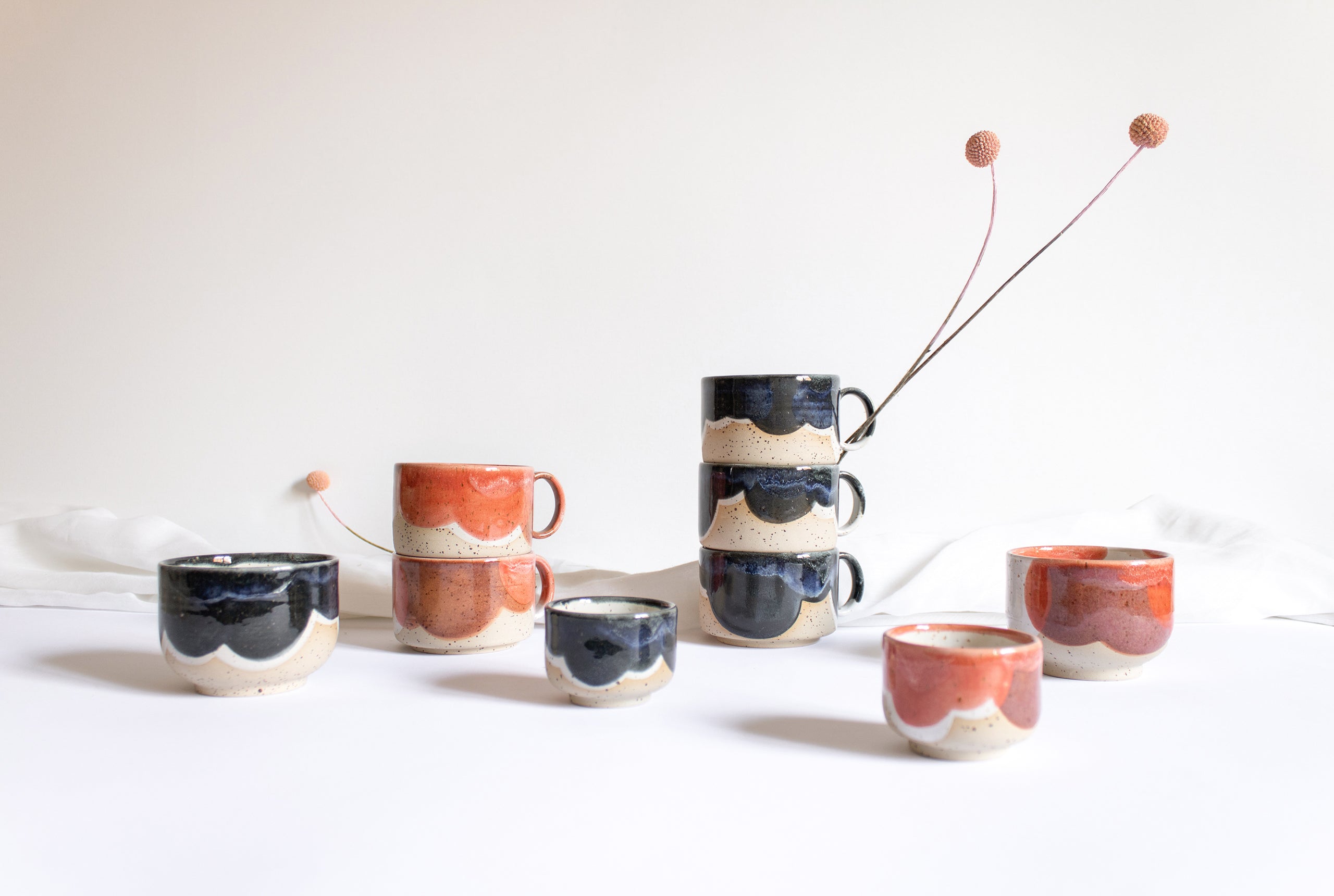 An assortment of artisanal tableware crafted from speckled cream clay and glazed in rich coral red and deep blue tones, arranged against white backdrop.