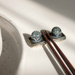 Handcrafted midnight blue chopstick rest with wooden chopsticks, placed next to a speckled white dining set.