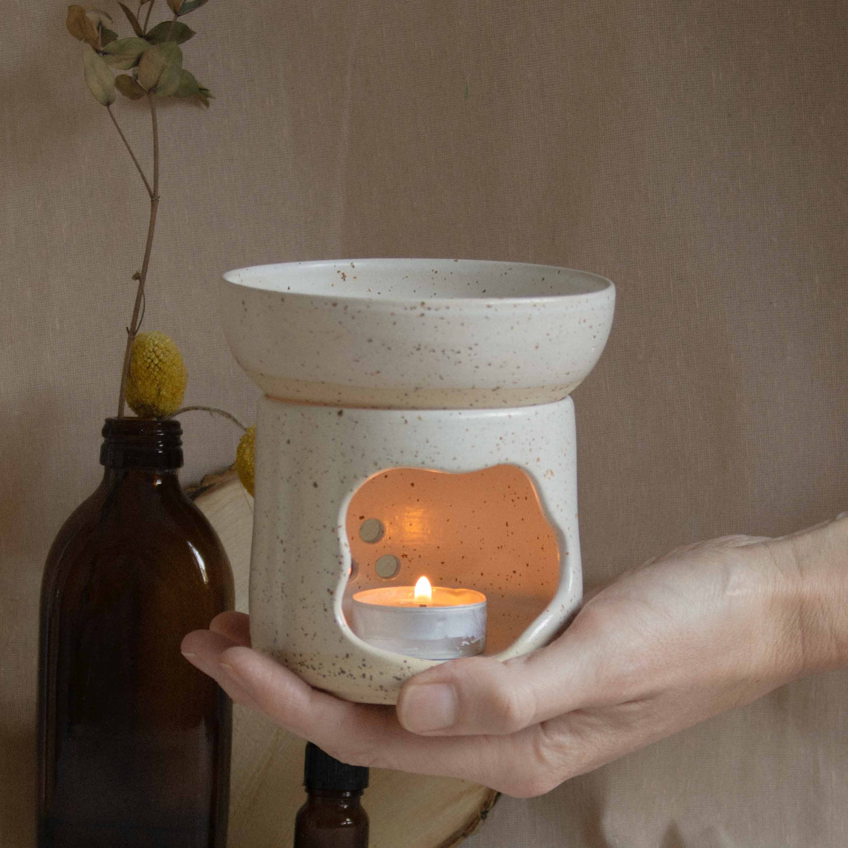 Hand holding a handmade oil burner in speckled white finish, with a lit candle inside, casting warm light.