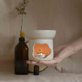 Hand holding a handmade oil burner in speckled white finish, with a lit candle inside, casting warm light.