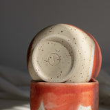 Close-up of a maker's mark detail at the bottom of a handmade ceramic cup crafted from speckled cream clay and coated in a coral red glaze.