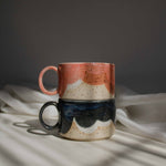 A stack of two handcrafted ceramic mugs in vivid coral red and deep blue tones, illuminated by a beam of light.