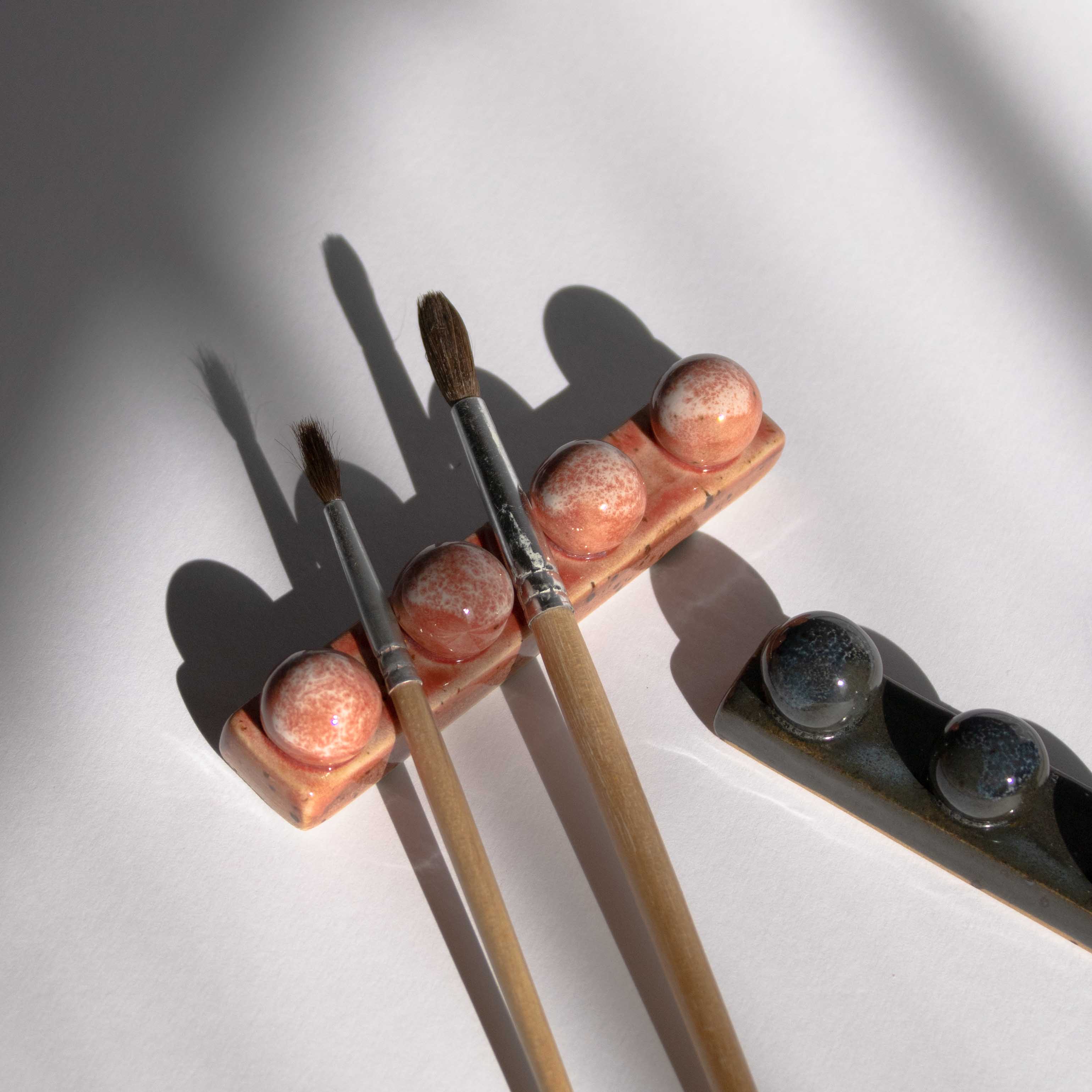 Ceramic paintbrush holder in coral red finish holding two thin watercolor brushes, with one deep blue paintbrush holder placed beside it.