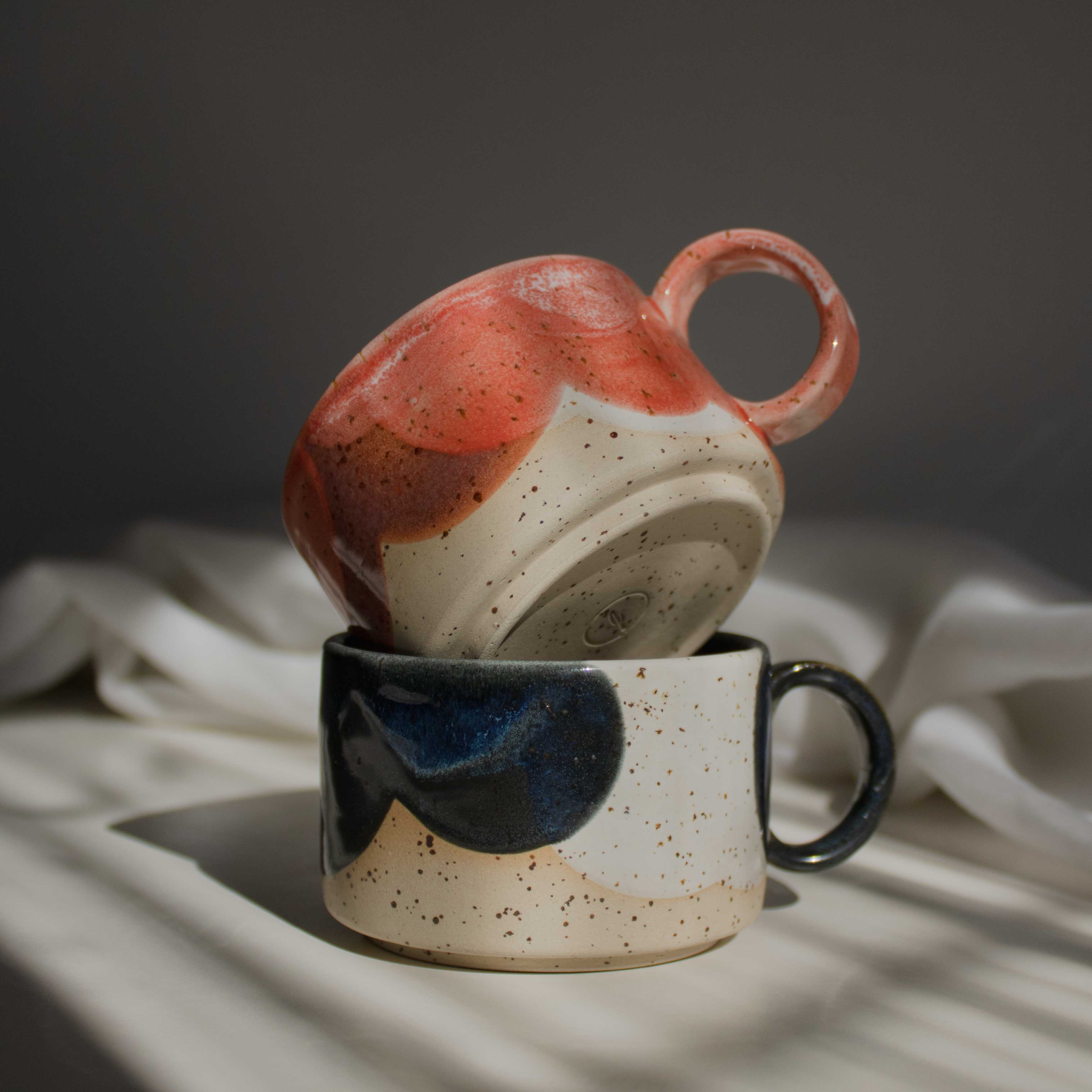 A stack of two handcrafted ceramic mugs in vivid coral red and deep blue tones, illuminated by a beam of light.