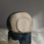 Close-up of a maker's mark detail at the bottom of a handmade ceramic cup crafted from speckled cream clay and coated in a deep blue glaze.