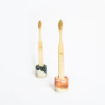 Two wooden toothbrushes displayed in coral red and blue handmade ceramic holders against a white background.