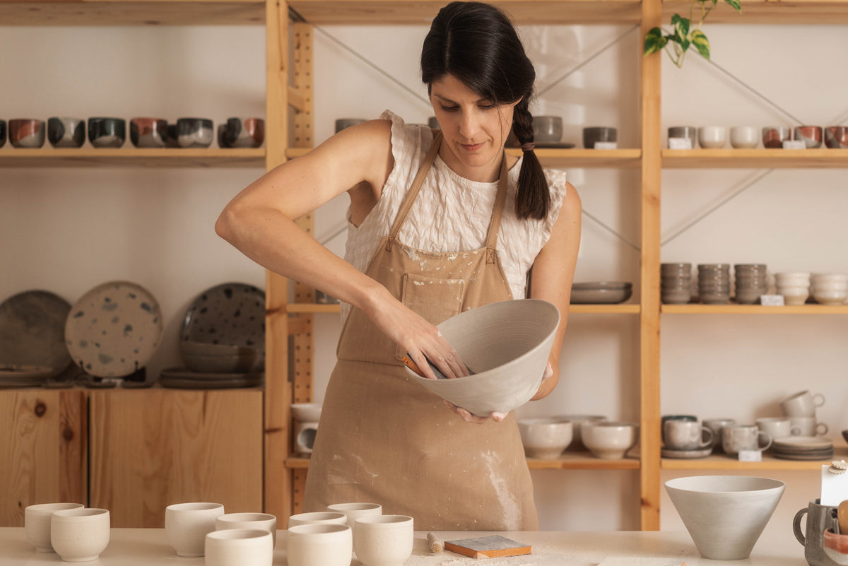 Potter standing in pottery studio, sanding a ceramic bowl on a worktable.