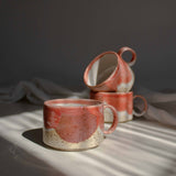 A group of multiple handcrafted ceramic mugs in vivid coral red tones, illuminated by a beam of light.