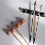 Overhead view of one deep blue and one coral red paintbrush holder, each holding paintbrushes, illuminated by gentle sunlight.