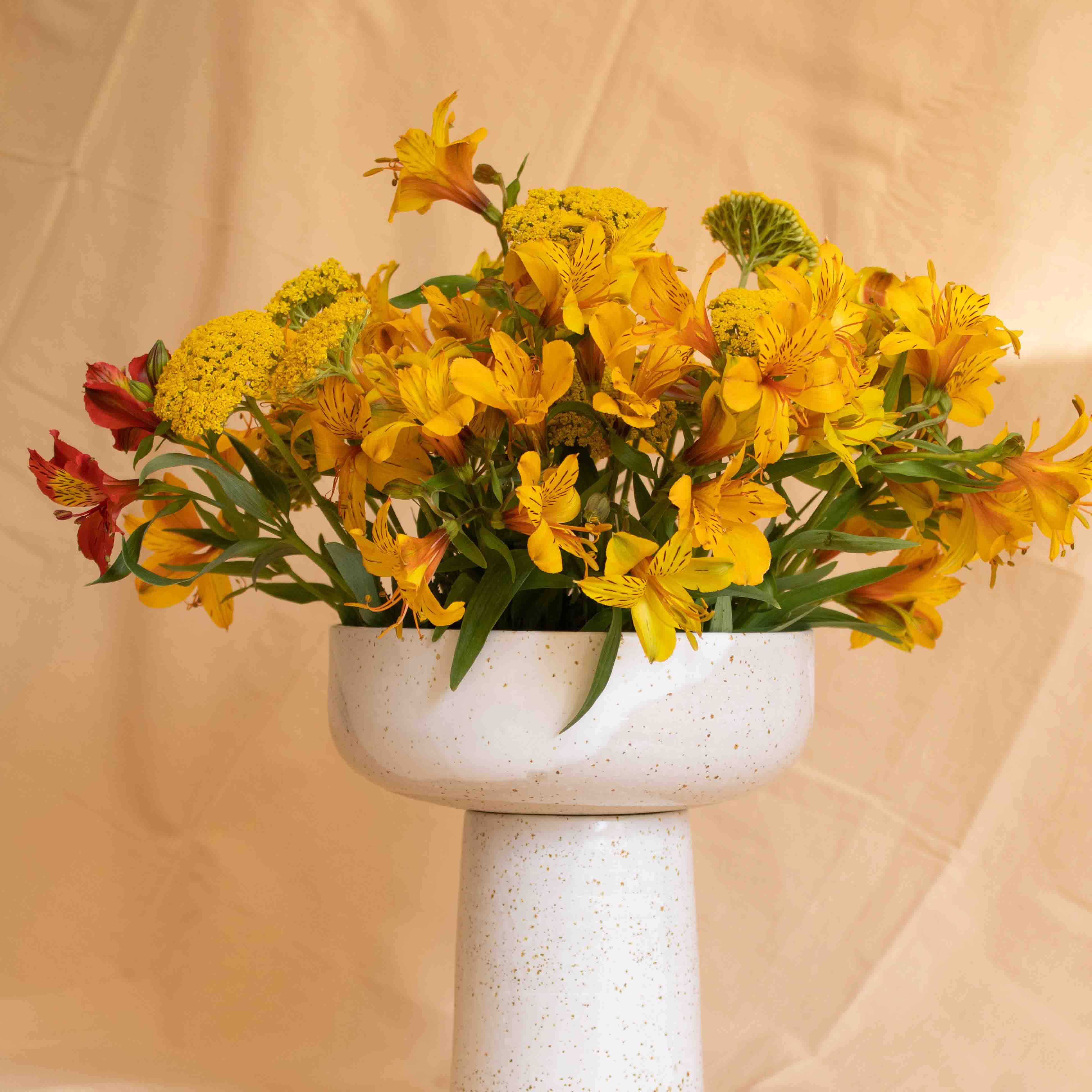 A handcrafted ceramic vase in speckled white finish, holding a bouquet of yellow flowers, set on an orange background.