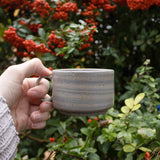 A hand holding a beautifully crafted ceramic mug, set against a backdrop of shrubs adorned with vibrant red fall berries.