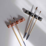 Overhead view of one deep blue and one coral red paintbrush holder, each holding paintbrushes, illuminated by gentle sunlight.