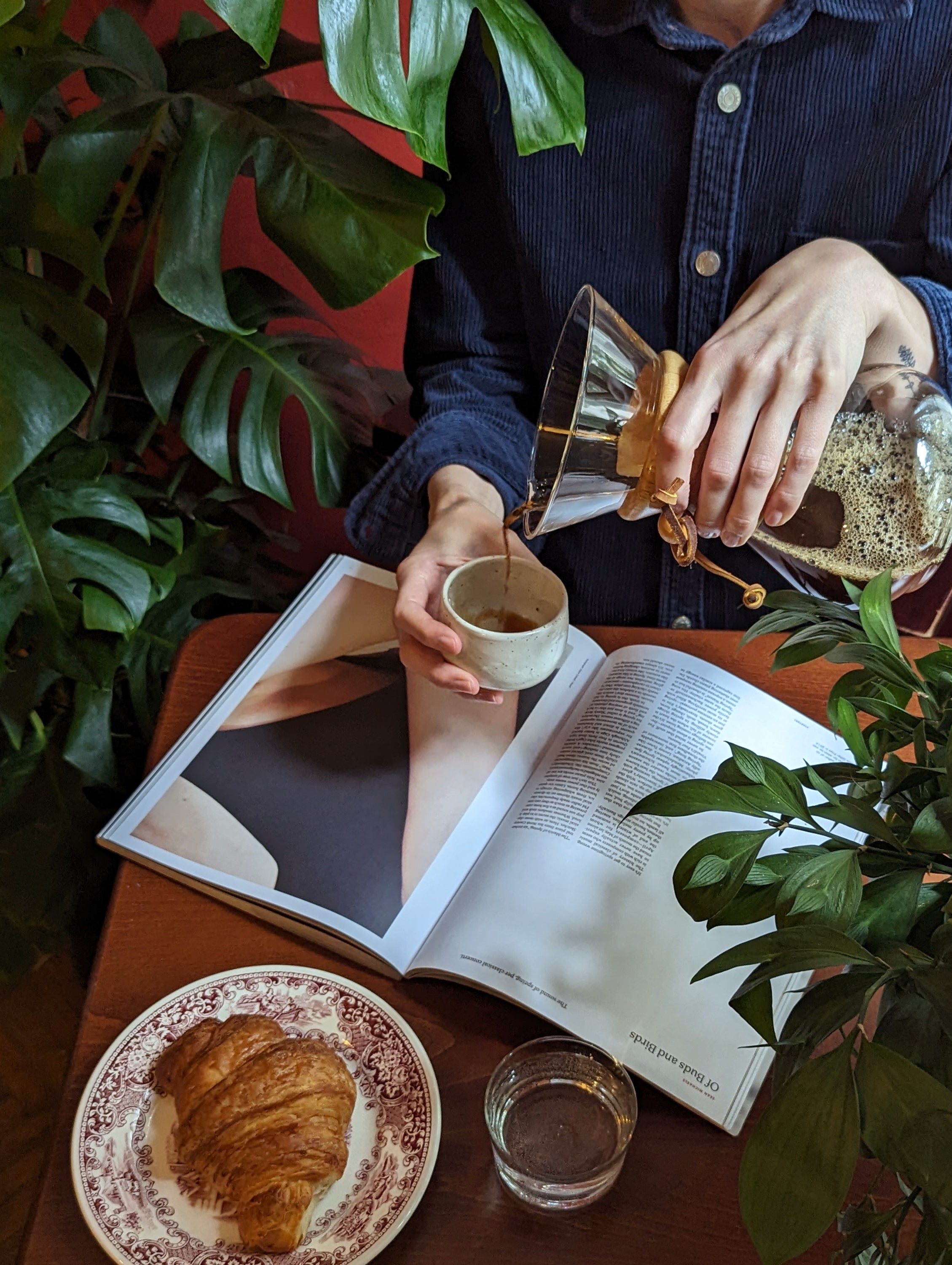 A person pours drip coffee into a speckled white ceramic cup, set on a tabletop adorned with magazines and croissants.