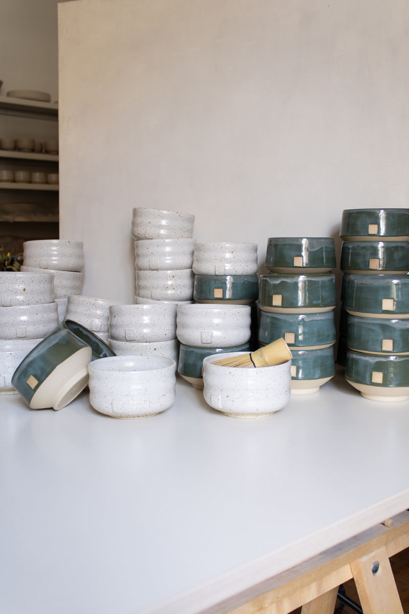 Multiple handmade matcha bowls, featuring green and white finishes, arranged in stacks on a white tabletop.