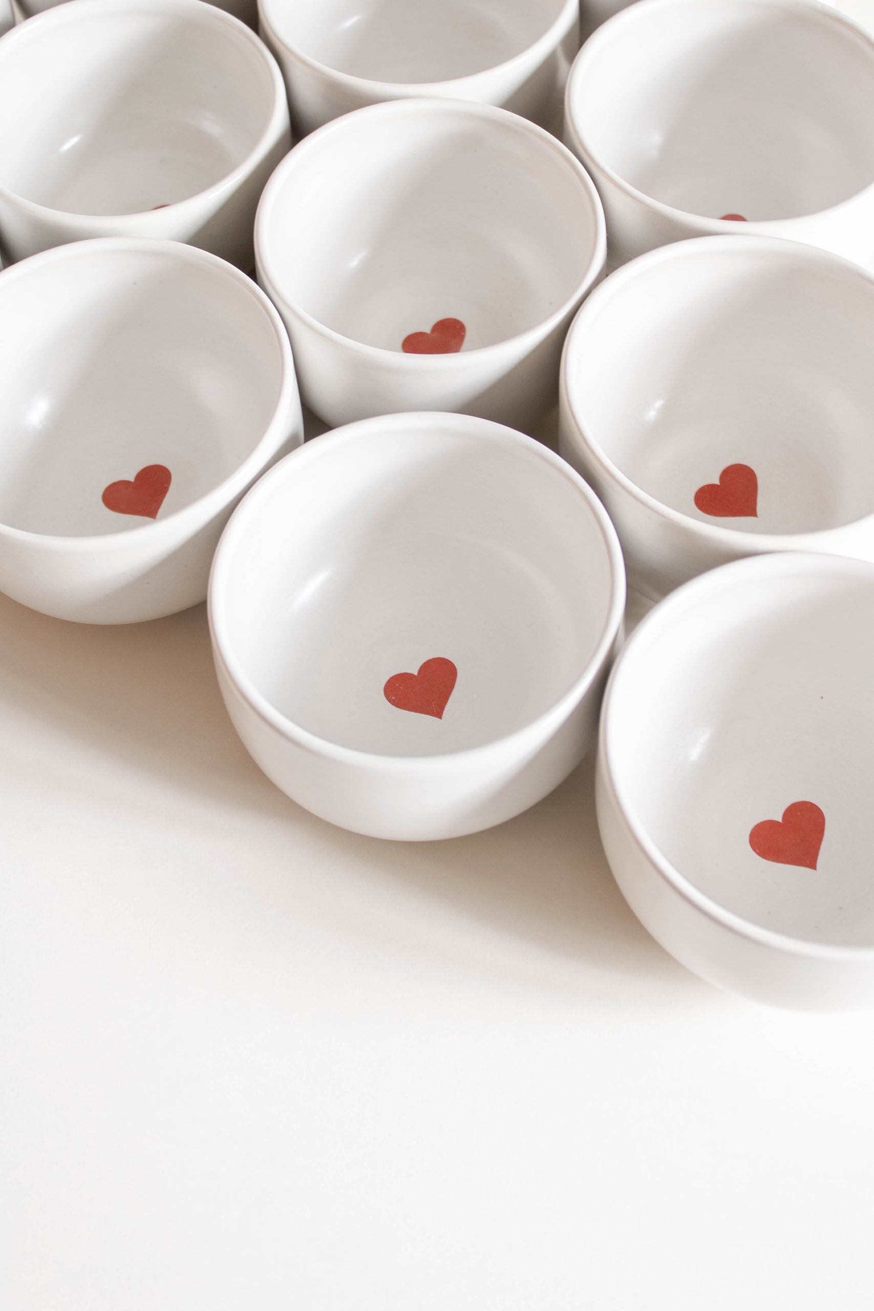 An array of white ceramic cups, featuring a small red heart positioned inside the bottom of each cup.