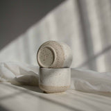 A stack of two handcrafted cappuccino cups in speckled white finish, with the top one featuring a maker's mark detail at the bottom of the cup.