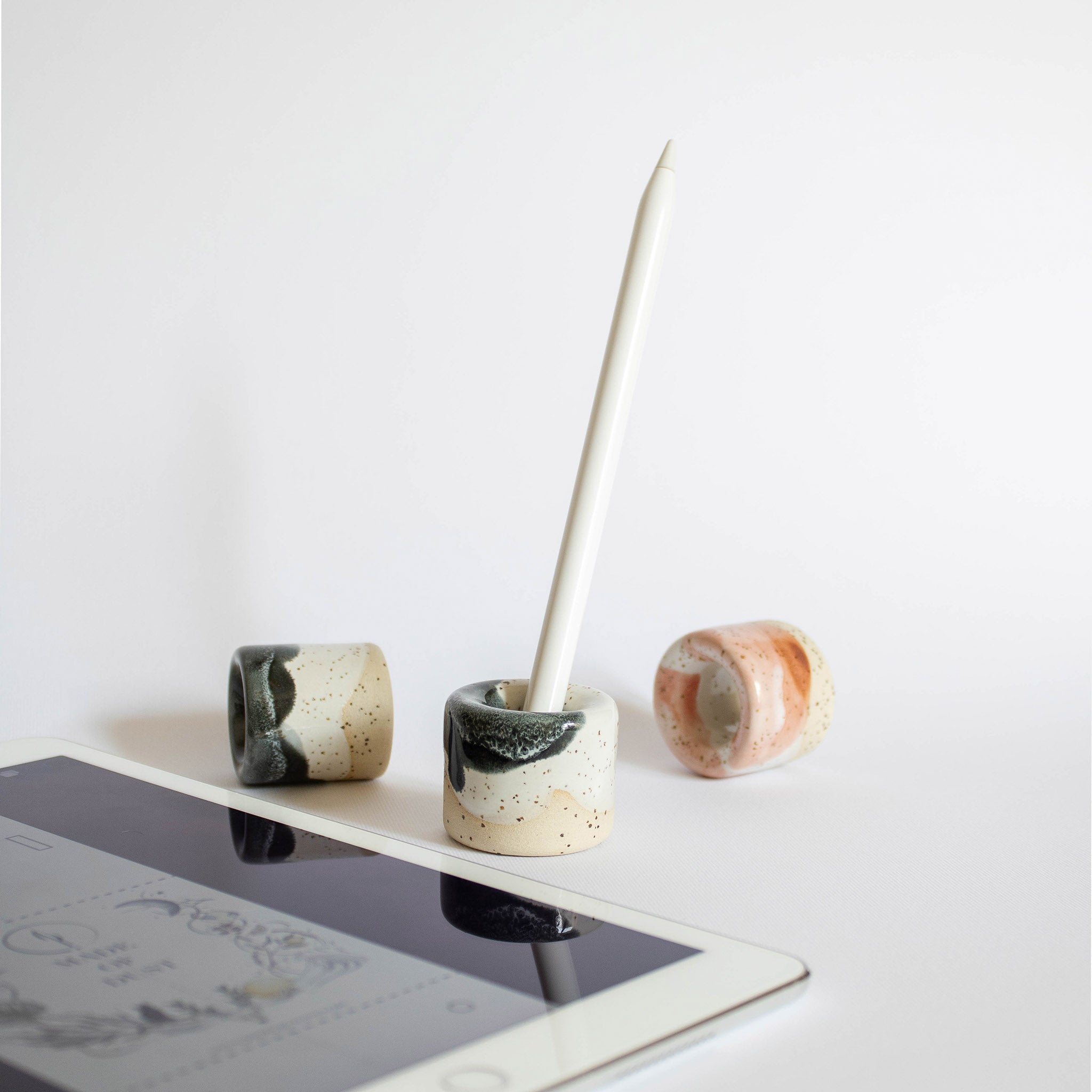 Multiple ceramic pen holders arranged next to an iPad, one holder contains an Apple Pencil, set against a white background.