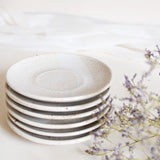 A stack of handcrafted ceramic saucer plates in speckled white finish, displayed on a white table cloth with dry flowers.