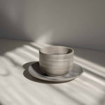 Handcrafted ceramic cup with a matte earthy finish, on a belonging saucer plate, in a beam of sunlight.