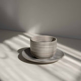 Handcrafted ceramic cup with a matte earthy finish, on a belonging saucer plate, in a beam of sunlight.