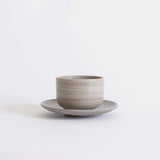 Handcrafted ceramic cup with a matte earthy finish, on a belonging saucer plate, displayed on a white background.