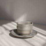 Handcrafted ceramic cup with a matte earthy finish, on a belonging saucer plate, in a beam of sunlight.