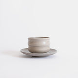 Handcrafted ceramic cup with a matte earthy finish, on a belonging saucer plate, displayed on a white background.