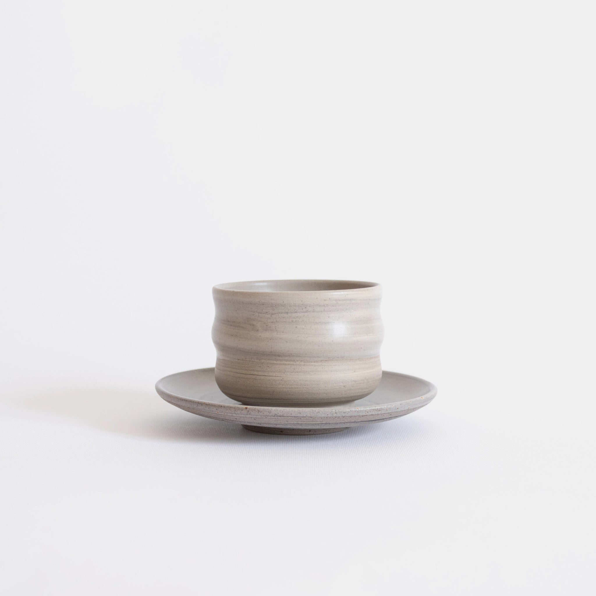 Handcrafted ceramic cup with a matte earthy finish, on a belonging saucer plate, displayed on a white background.