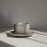 Handcrafted ceramic cup with a matte earthy finish, on a belonging saucer plate, in a beam of sunlight.