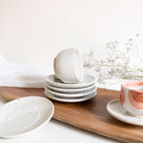 Handmade speckled white ceramic cappuccino cup with saucer plates on wooden board, with a blurred floral branch in the background.