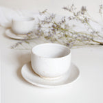 A handcrafted ceramic cup and saucer plate in speckled white finish, displayed with white table cloth and dry flowers.