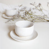 A handcrafted ceramic cup and saucer plate in speckled white finish, displayed with white table cloth and dry flowers.