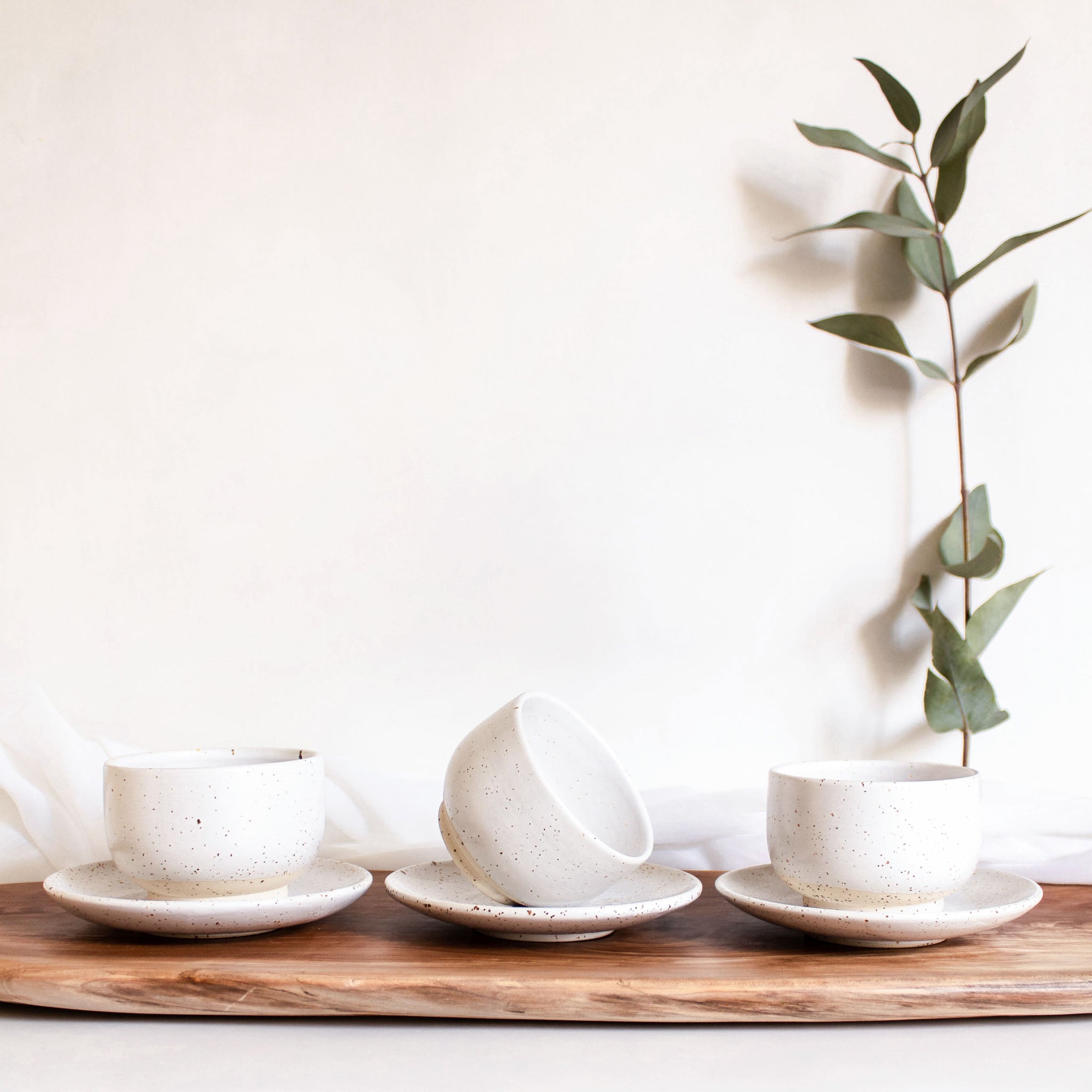 A linear arrangement of three handcrafted ceramic Pure cups with belonging saucer plates in speckled white finish, displayed on a wooden board.