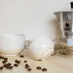 Speckled white handmade espresso cup resting on a wooden table beside a cappuccino cup, a metal Bialetti moka pot, and scattered coffee beans.