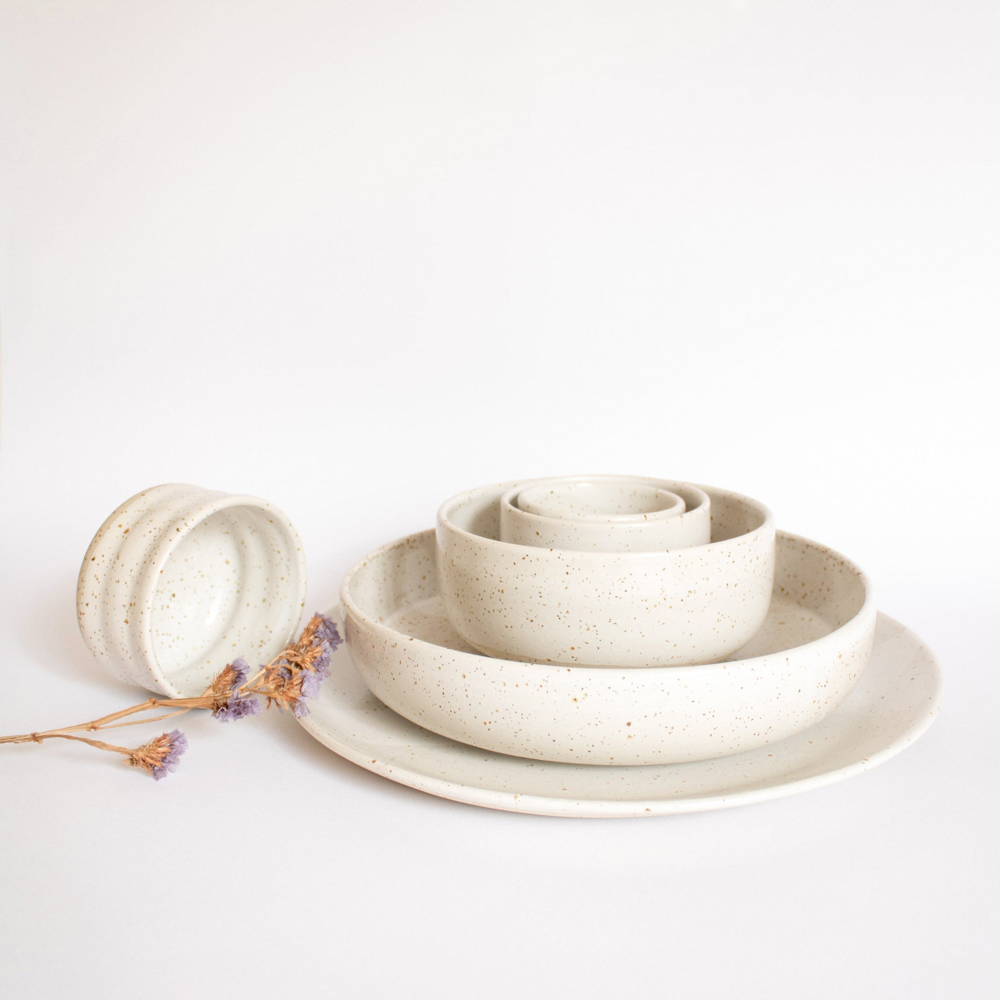 Handcrafted dining set in speckled white finish, featuring plates, bowls, and cups arranged on a white surface.