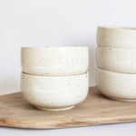 Handcrafted ceramic bowls in speckled white finish arranged on a rustic wooden board.