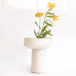 A handcrafted ceramic vase in speckled white finish, holding flowers, set on a white background.