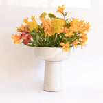 A handcrafted ceramic vase in speckled white finish, holding a bouquet of yellow flowers, set on a white background.