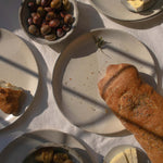 Handmade ceramic plate in speckled white finish, displayed with a selection of appetizers including olives, cheese, bread, and pears.