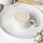 Close-up view of the base of a handcrafted ceramic cup with a handle, positioned upside-down on a speckled white plate.