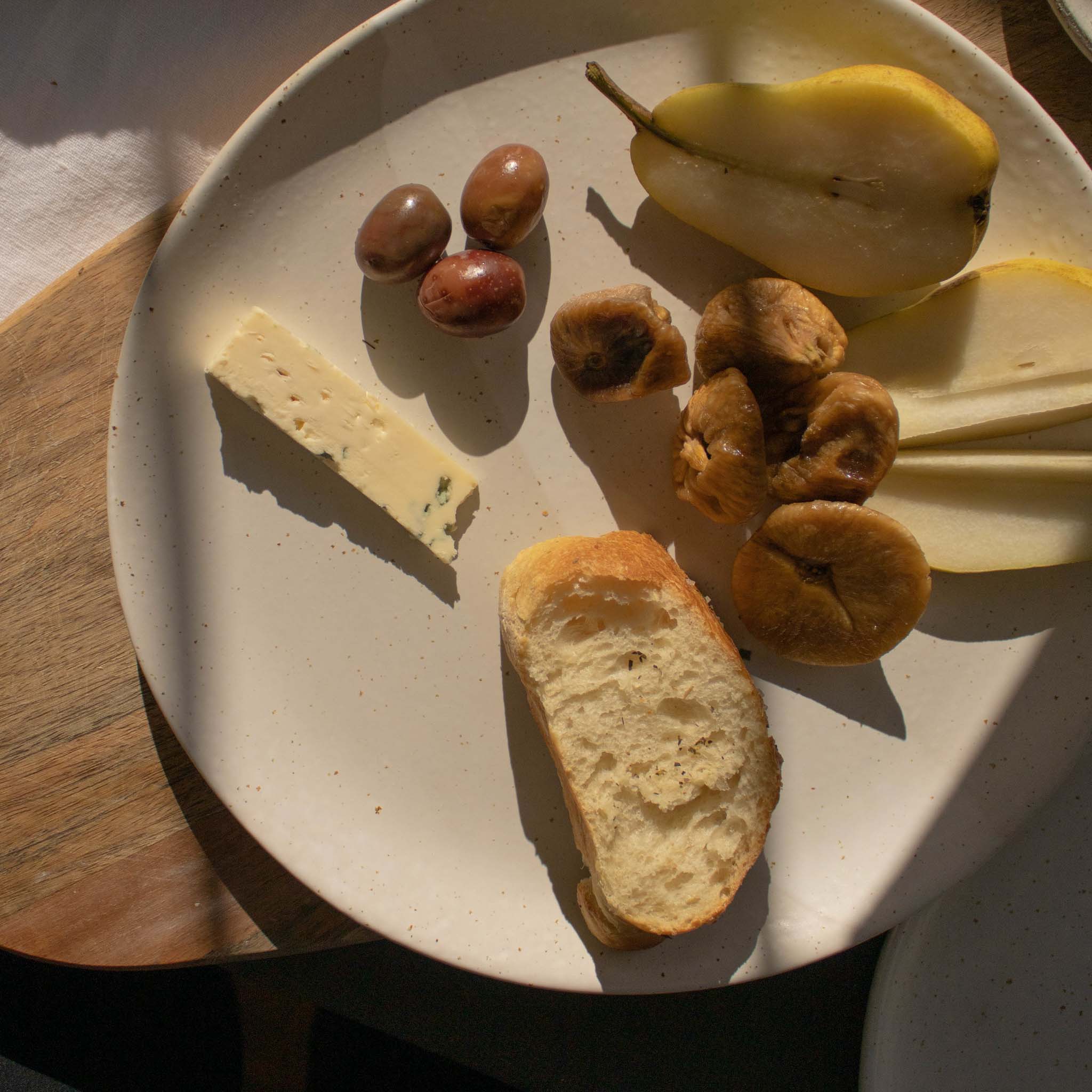 Handmade ceramic plate in speckled white finish, displayed with a selection of appetizers including olives, cheese, bread, and pears.