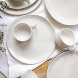 Artfully scattered on a table, speckled white handmade ceramic cups with handles rest atop matching speckled white plates.