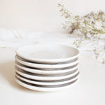 A stack of handcrafted ceramic saucer plates in speckled white finish, displayed on a white table cloth with dry flowers.