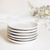 A stack of handcrafted ceramic saucer plates in speckled white finish, displayed on a white table cloth with dry flowers.