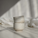 A stack of two handcrafted espresso cups in speckled white finish, illuminated by contrasting light and shadows.