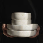 Handcrafted white speckled plates and bowls held in hands against a black backdrop, illuminated by sunlight.