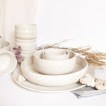 Handcrafted dining set in speckled white finish, featuring plates, bowls, and cups arranged on a white surface.