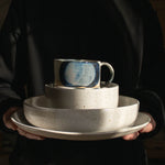 Hands holding handcrafted speckled white plates, a bowl, and a blue cup against a black backdrop, bathed in sunlight.