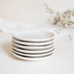 A stack of handcrafted ceramic saucer plates in speckled white finish, displayed on a white table cloth with dry flowers.