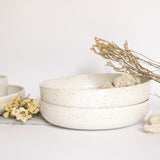 Handcrafted ceramic pasta plates in speckled white finish, displayed alongside rocks and branches on a white background.